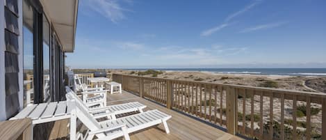 A wooden deck with white lounge chairs and a table, overlooking a beach with dunes and the ocean.