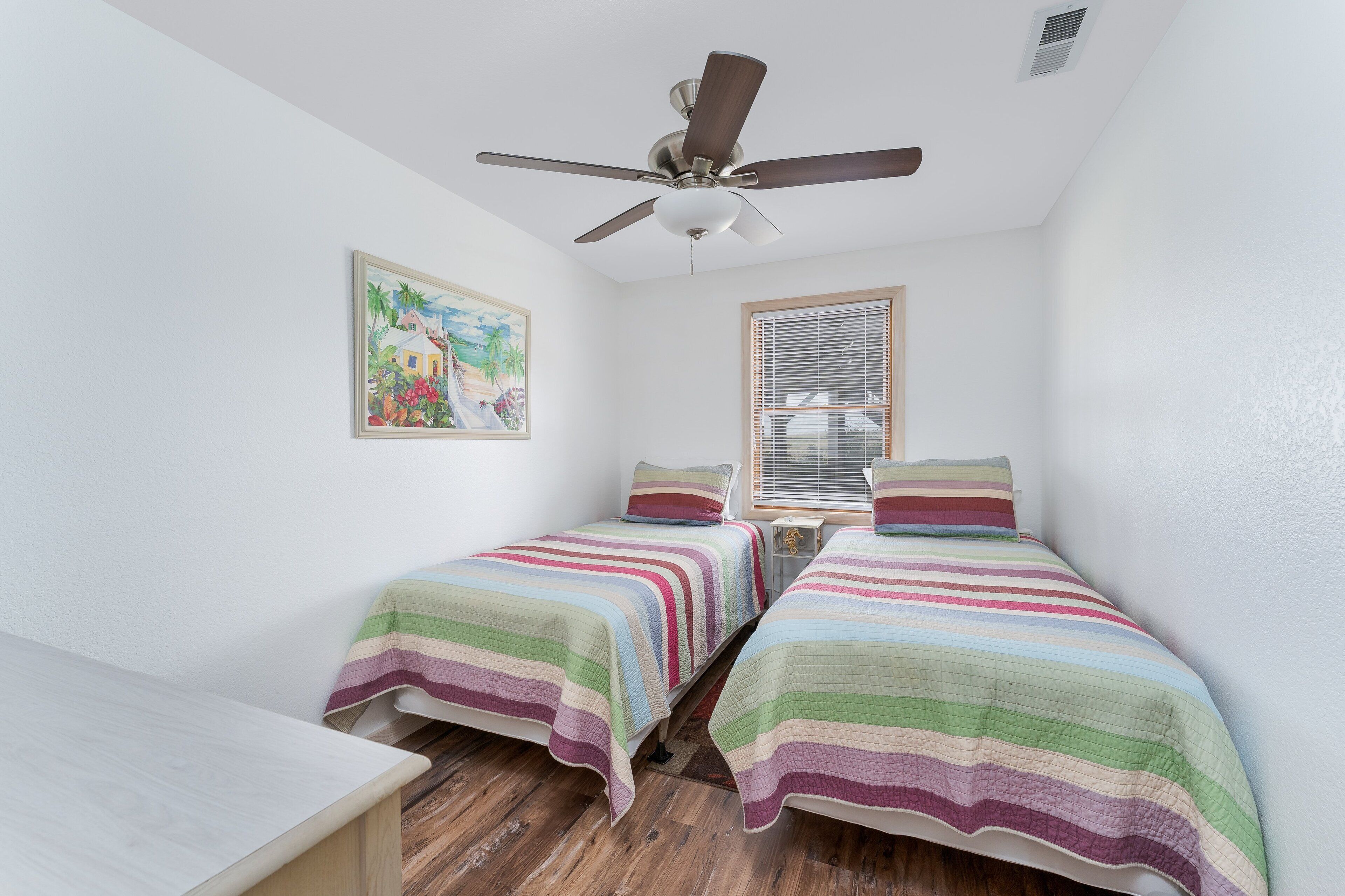 A small, clean room with two single beds, a ceiling fan, a window with blinds, and a painting on the wall.