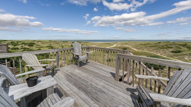 Een houten terras met Adirondack-stoelen en een emmer, met uitzicht op een strand en duinen.