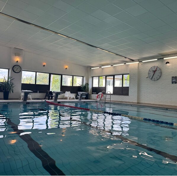 An indoor swimming pool with clear lanes, a clock, and windows with a view of trees.