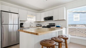 Una cocina moderna con armarios blancos, electrodomésticos de acero inoxidable, una isla de madera con taburetes y un amplio ventanal con persianas.