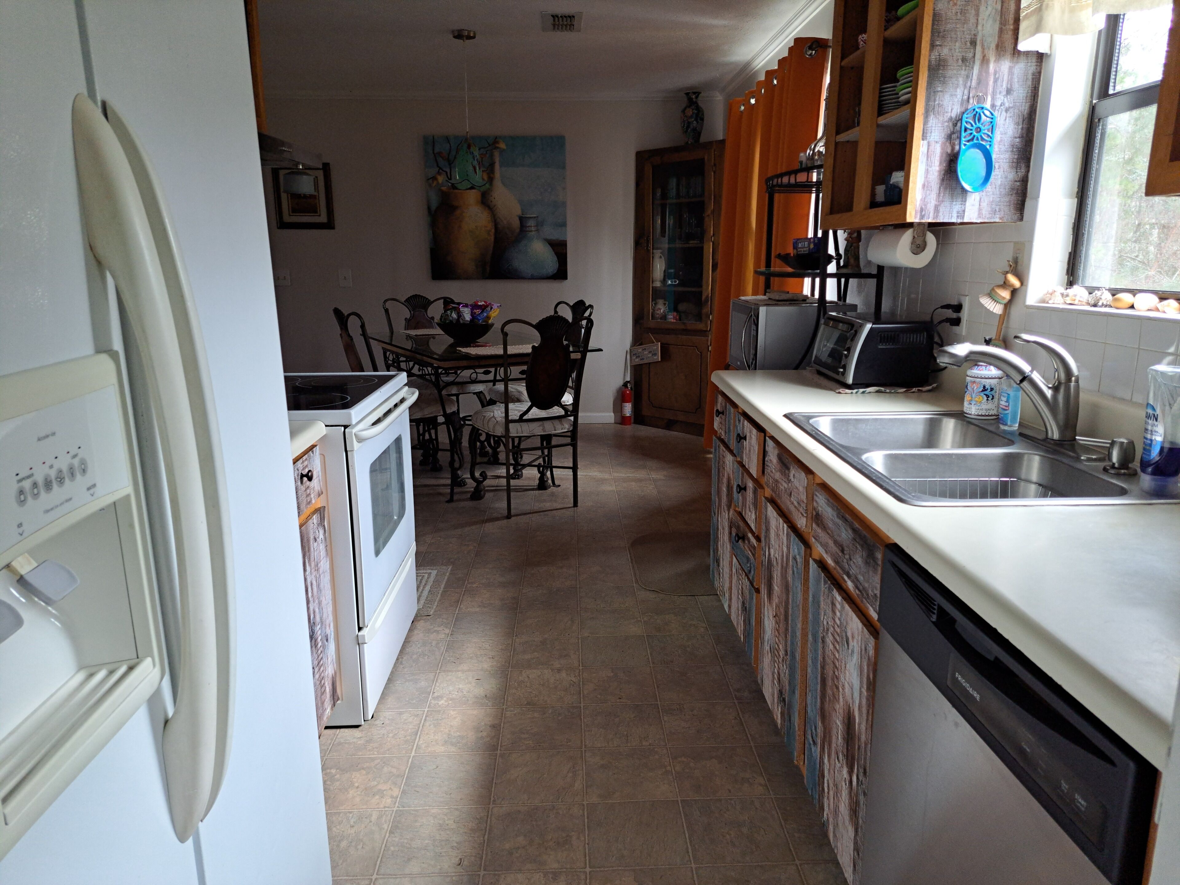 Uma cozinha com fogão, forno, pia e armários. Há uma mesa de jantar com cadeiras e um quadro na parede.