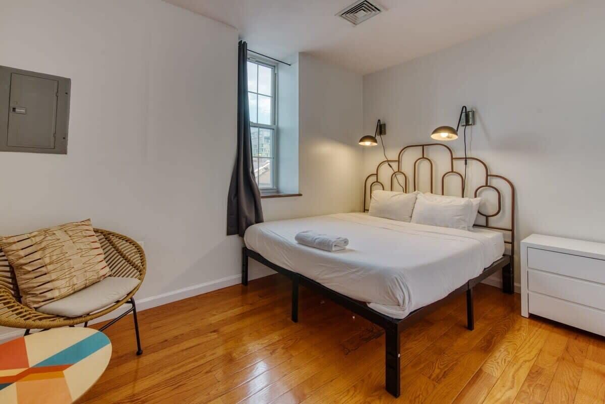 A bedroom with a bed, a chair, a bedside table, a lamp, and a window.