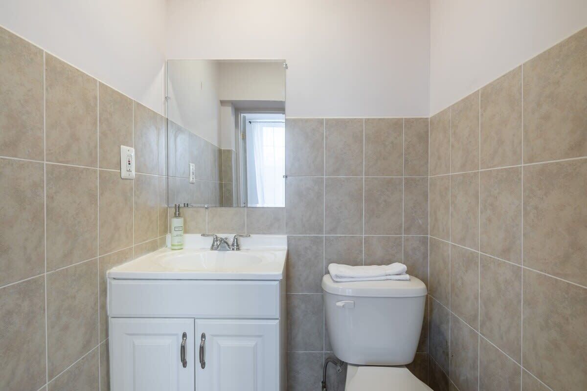 Une salle de bain avec un lavabo blanc, un miroir et des toilettes, dont le sol et les murs sont recouverts de carreaux de couleur beige clair.