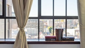Een raam met uitzicht op een stadsbeeld, een gordijn omhoog geknoopt, en een stapel boeken op de vensterbank.