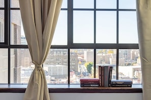 A window with a view of a cityscape, a curtain tied at the top, and a stack of books on the windowsill.