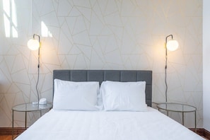 Una cama bien hecha con dos almohadas, un cabecero tapizado en gris y dos mesitas de noche con lámparas.