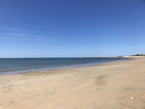 A wide sandy beach with gentle waves, a clear sky, and a distant shoreline with sparse vegetation.