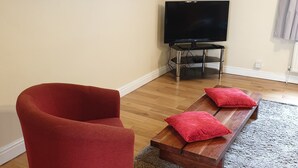 Un salon avec un fauteuil rouge, un banc en bois avec des coussins rouges, un téléviseur à écran plat et un grand tableau floral accroché au mur.