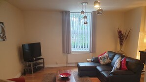 Un salon avec un canapé en cuir noir, une table basse en bois et un fauteuil rouge. On y trouve un téléviseur posé sur un meuble et une fenêtre avec des rideaux.