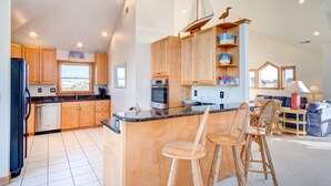 A modern kitchen with wooden cabinets, a central island with bar stools, and a dining area with a sofa and table. - 4052 - Mirth N Mist II - Spacious Duck Escape | Pool & Sleeps 17 (Duck)