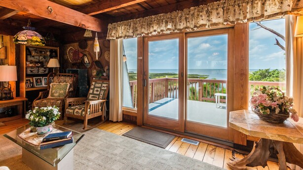 Ein gemütliches Cabin-Interieur mit Holzboden, Kamin, Essbereich und einer großen Schiebeglastür, die auf eine Terrasse mit Meerblick führt.
