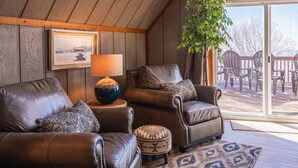 A cozy living room with leather sofas, a patterned rug, and a large window offering a view of outdoor furniture. - 6BR Mtn House, Spectacular Valley and Sunset Views, Rec Room, Hot Tub, Fire Pit (Snowshoe)