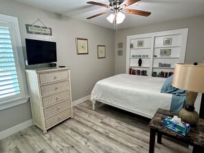 Un dormitorio con cama, cómoda, ventilador de techo, televisor y mesita de noche con lámpara.