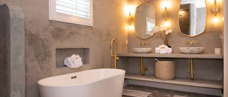 Un baño moderno con una bañera independiente, dos lavamanos con espejos sobre encimera y una ventana con persianas.