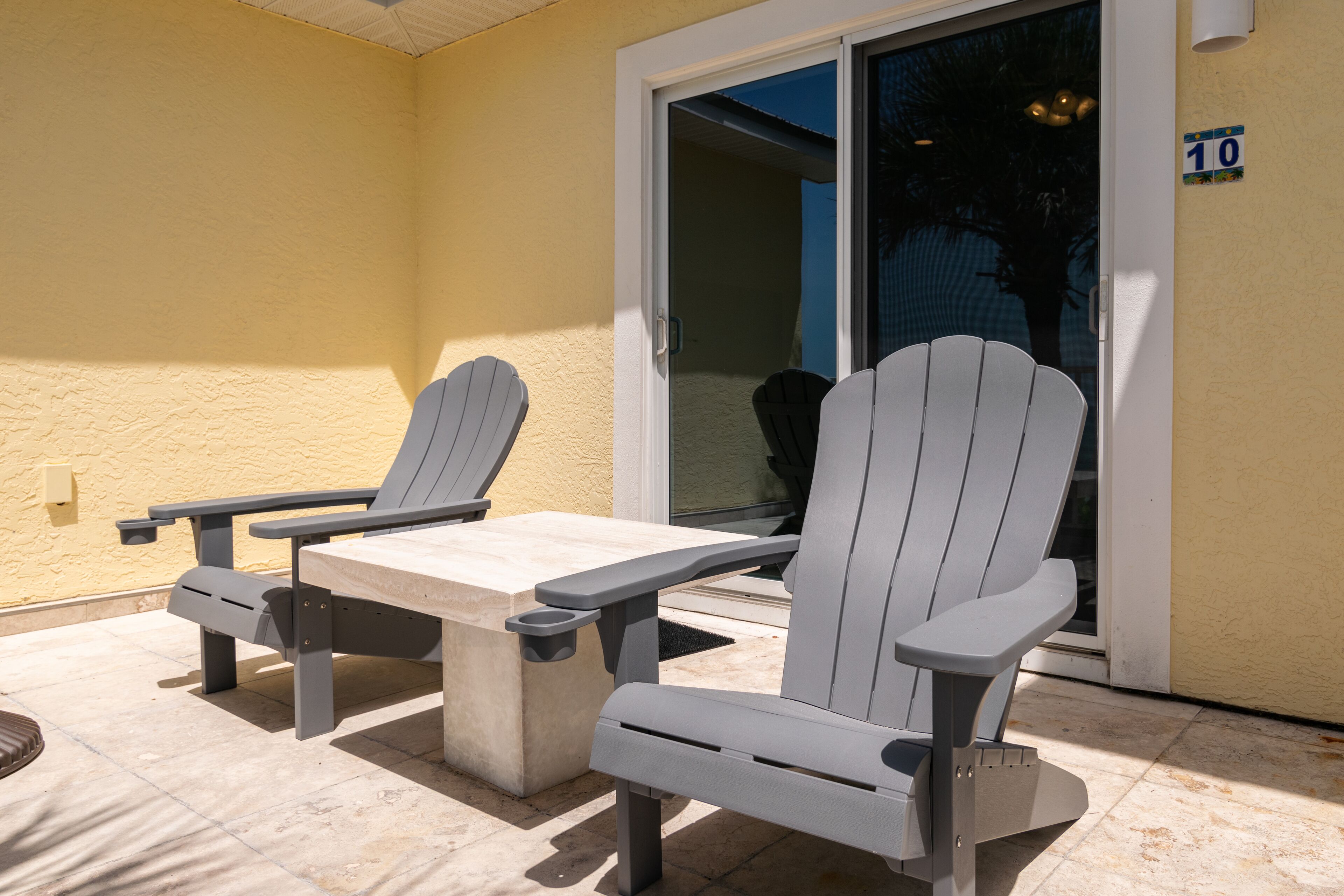 Two Adirondack chairs with a table on a patio with a yellow wall and a sliding glass door.