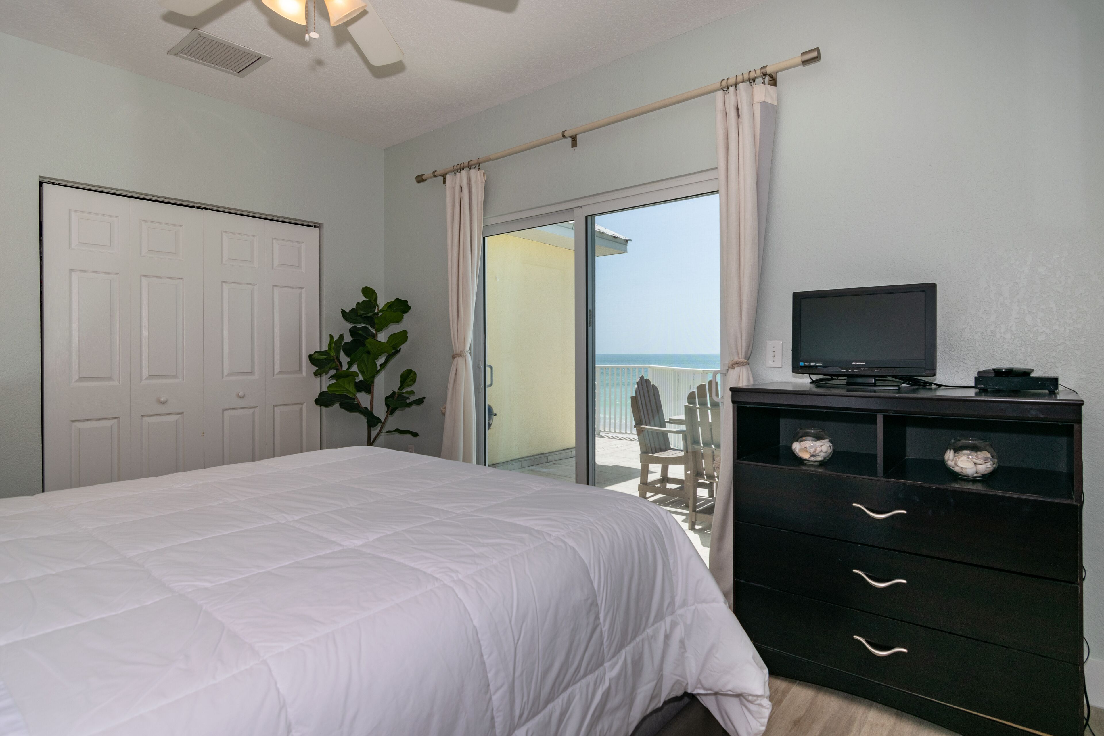 A bedroom with a bed, a TV on a dresser, and a view of the ocean through a sliding glass door.
