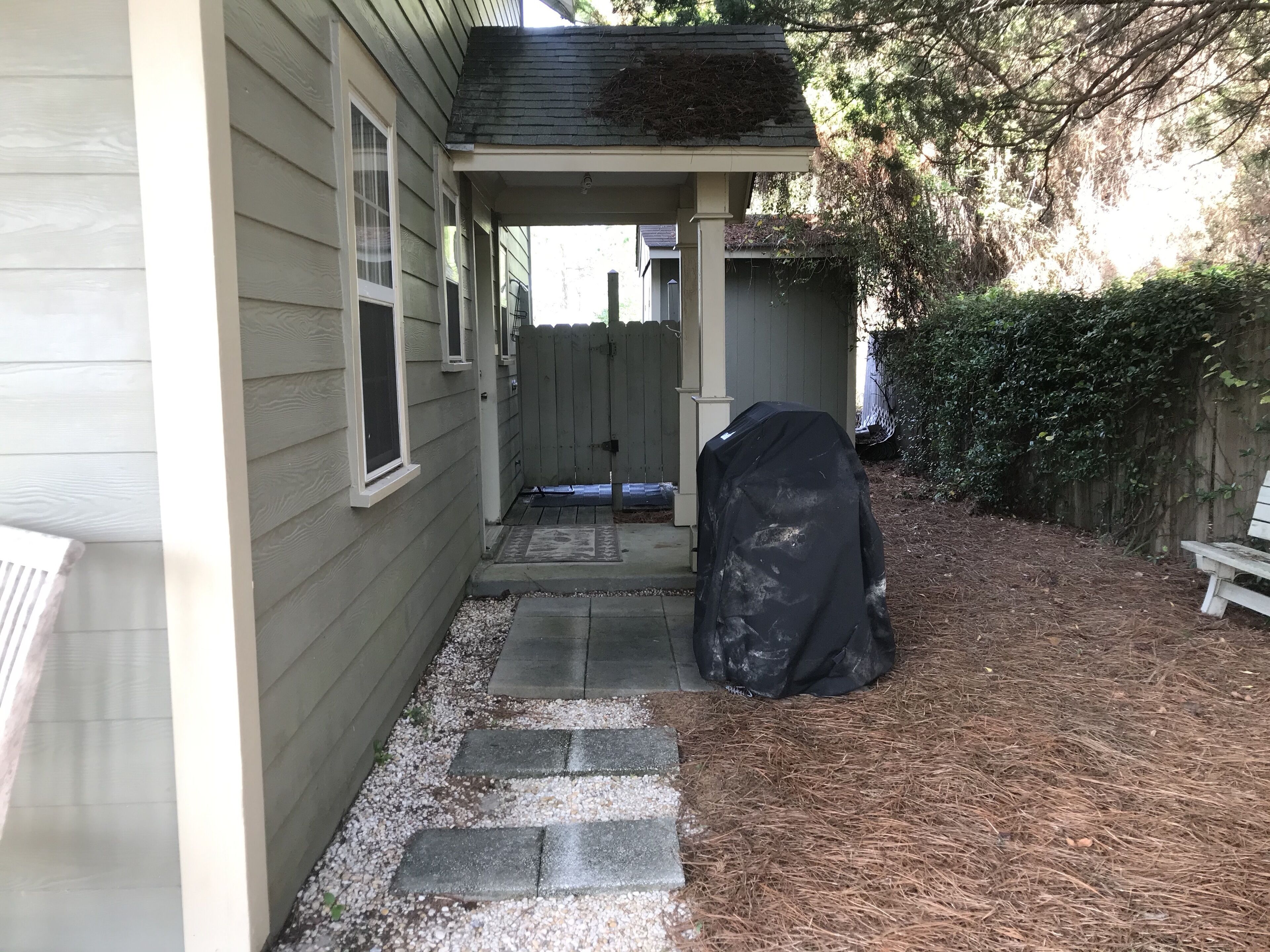 A covered porch with a wooden fence, a covered grill, and a gravel pathway leading to a house.