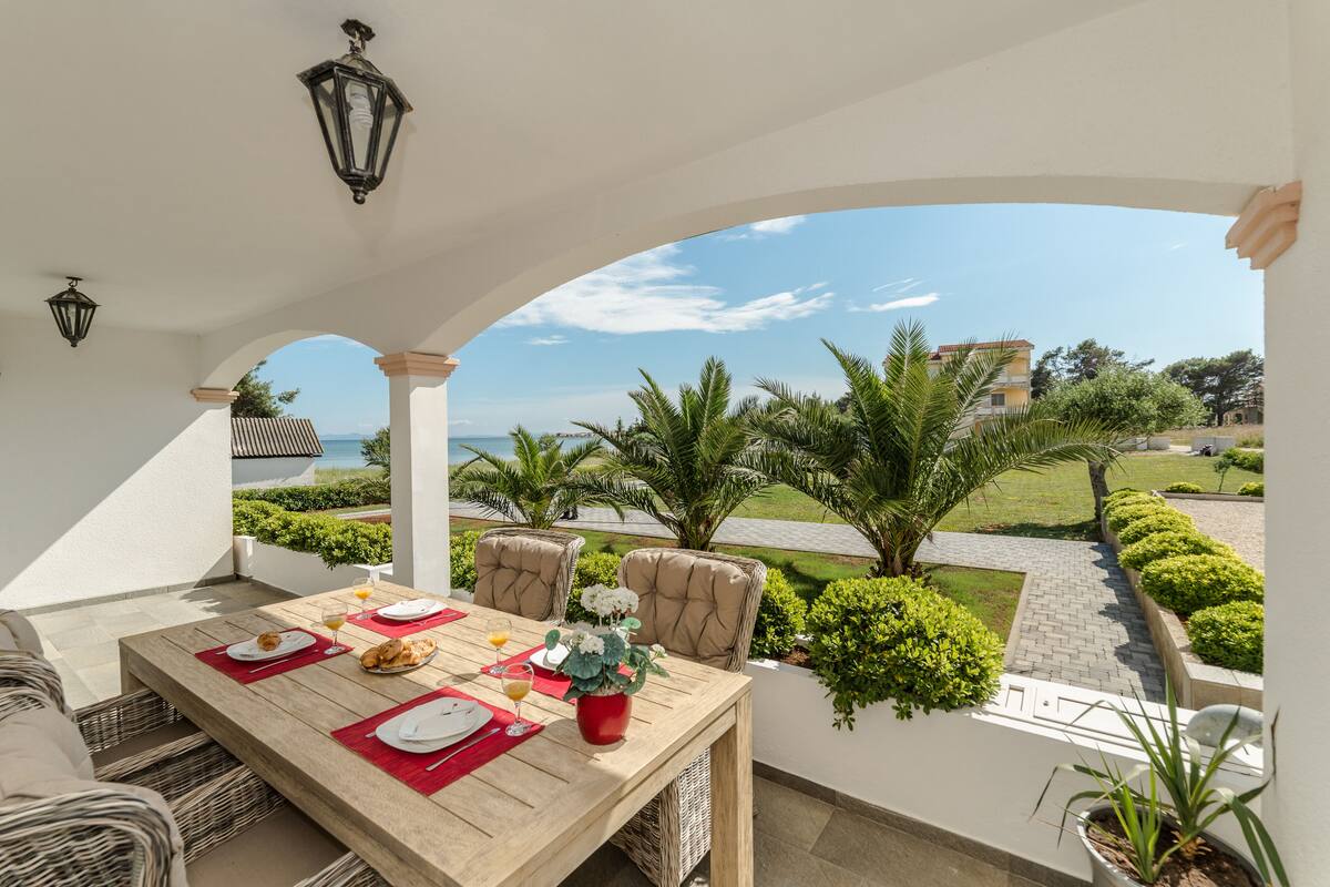 Un espace de restauration en plein air avec vue sur la mer, une table dressée avec des assiettes, des verres et une plante en pot.