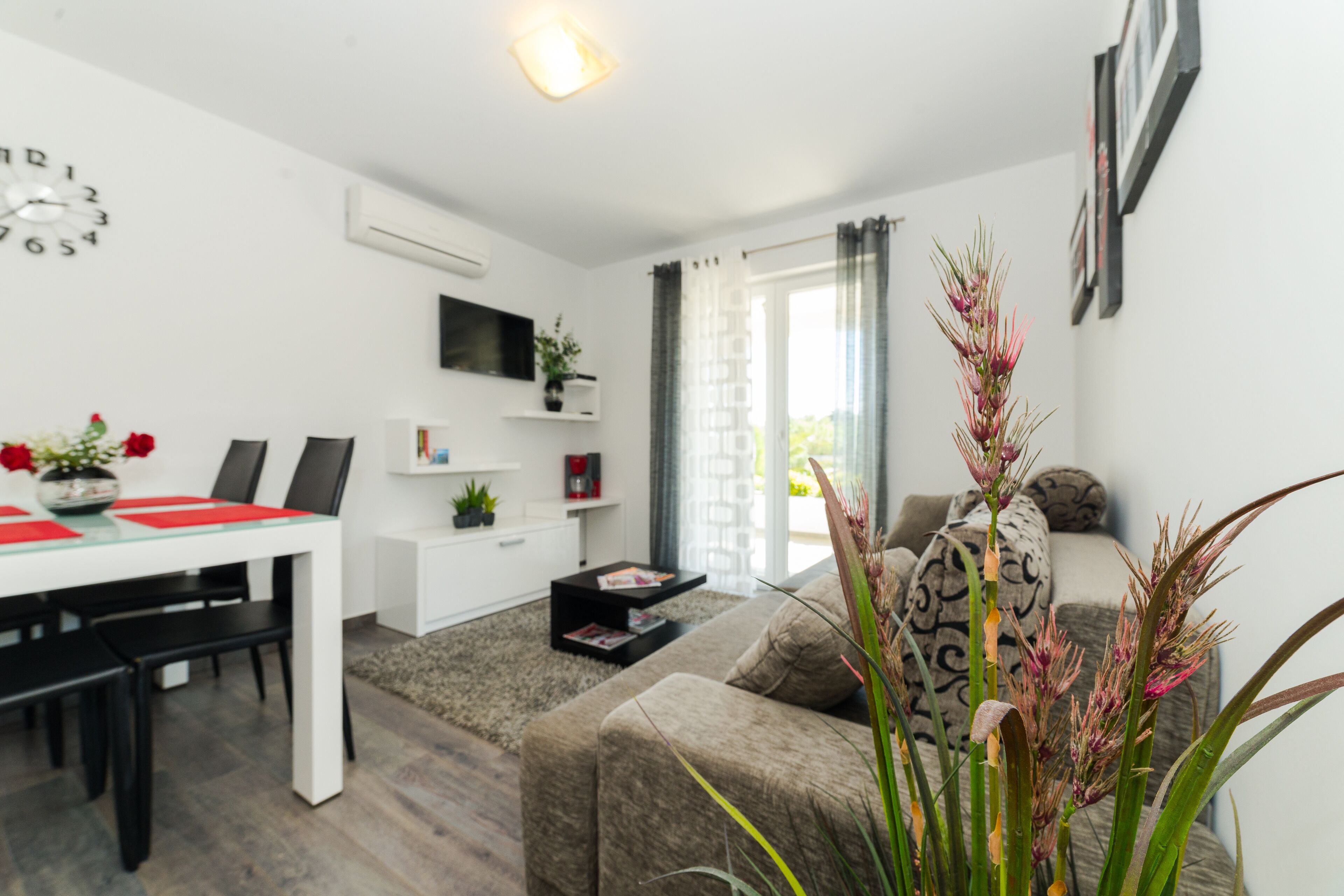 Un salon moderne comprenant un coin repas, un téléviseur à écran plat fixé au mur et une grande fenêtre avec des rideaux.