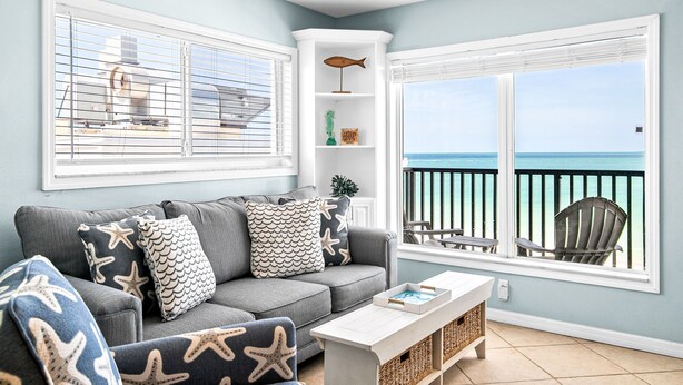 Un salon avec un canapé gris, des coussins à motifs étoilés, une table basse blanche, une petite étagère blanche et une vue sur l’océan par une porte-fenêtre donnant sur le balcon.