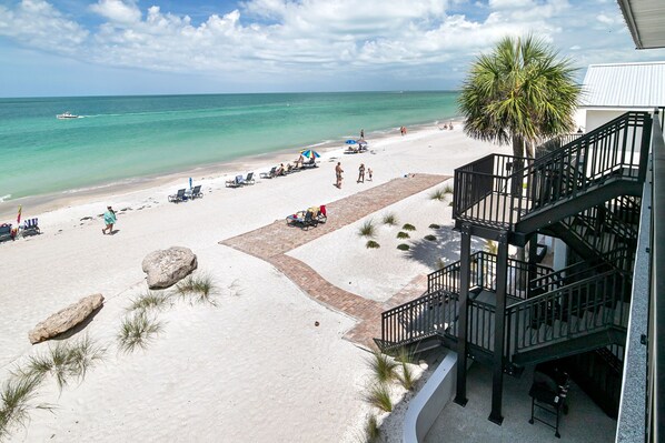 A beach view with a staircase leading to a balcony, lounge chairs, and a palm tree.
