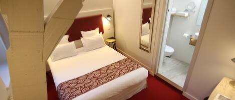 Une chambre d’hôtel avec un lit, une table de chevet, un miroir et une salle de bain visible à travers une porte ouverte.