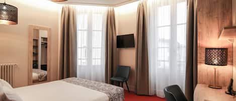 Una habitación de hotel con cama, escritorio, silla, televisor y ventanas grandes con cortinas.