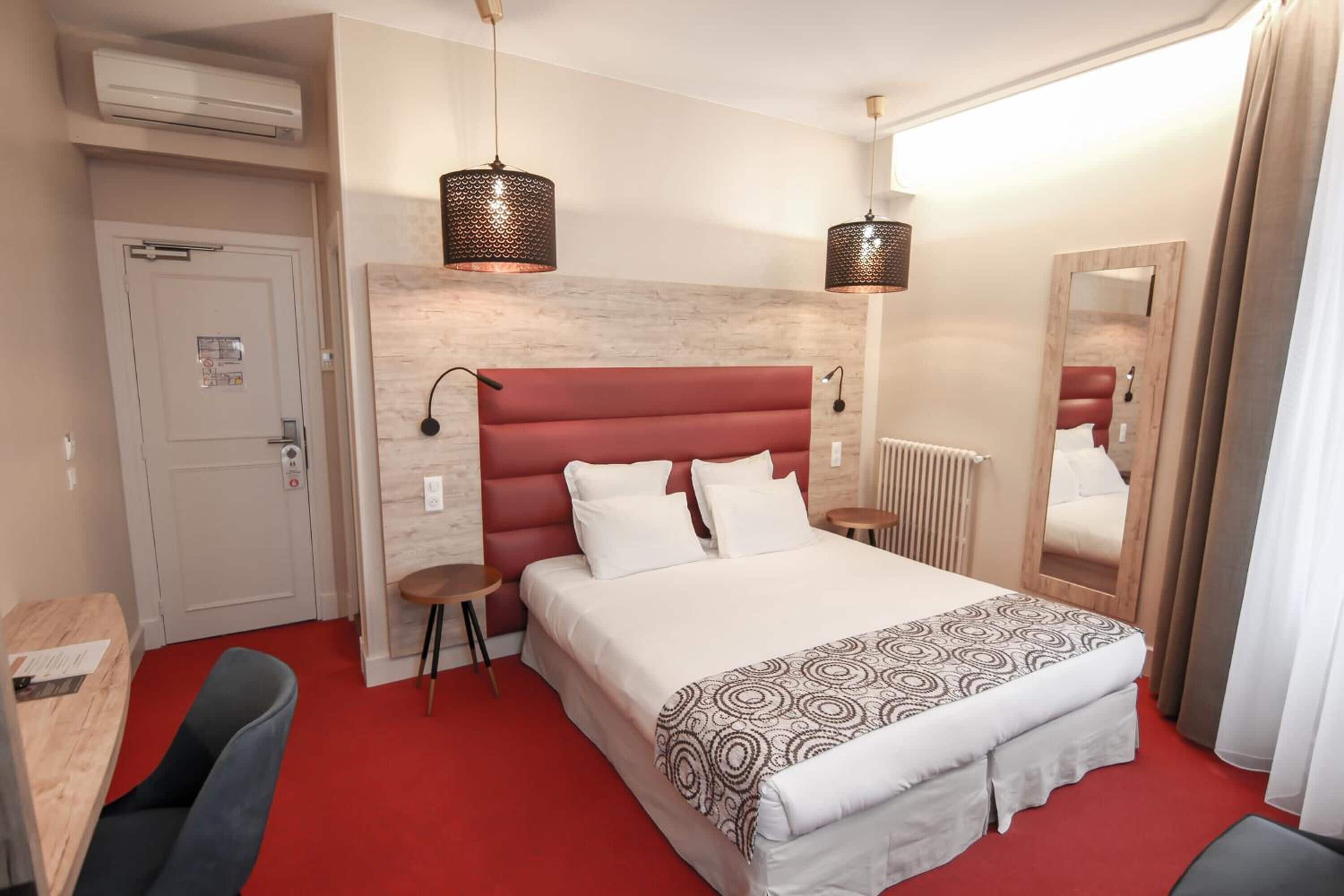 Une chambre d’hôtel avec un tapis rouge, un lit recouvert d’une couvre-lit à motifs, deux lampes de chevet, une tête de lit en bois, un miroir, un radiateur et un bureau avec une chaise.
