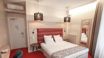 Une chambre d’hôtel avec un tapis rouge, un lit recouvert d’une couvre-lit à motifs, deux lampes de chevet, une tête de lit en bois, un miroir, un radiateur et un bureau avec une chaise.