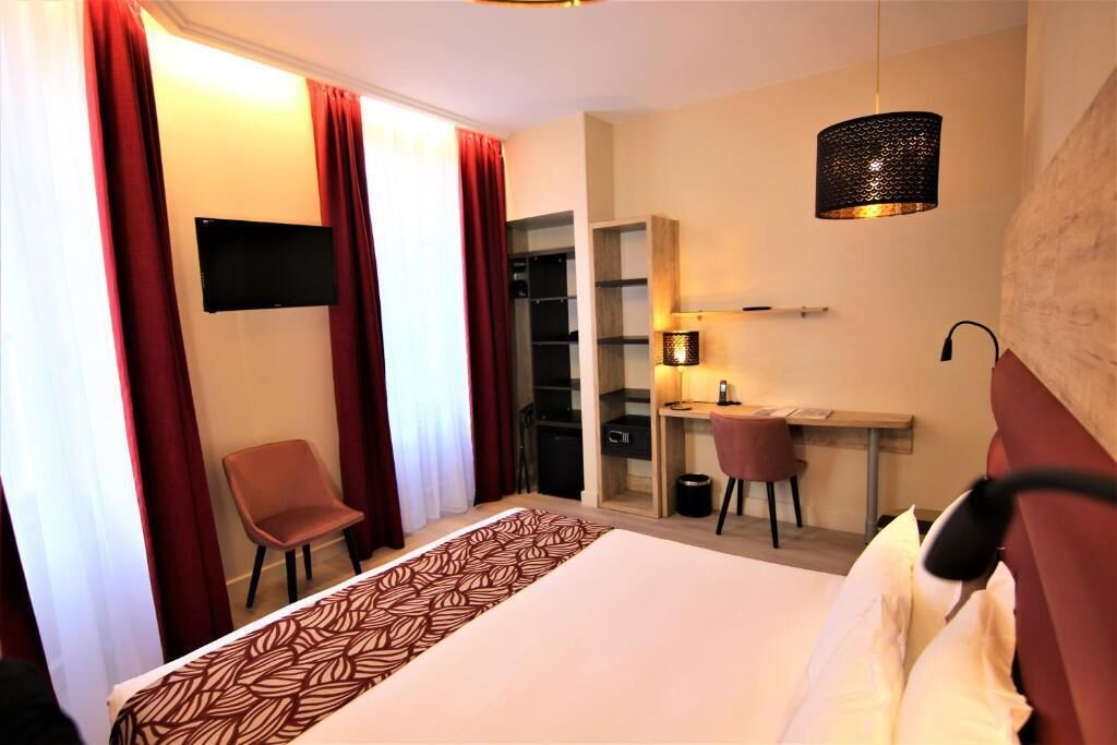 Quarto de hotel com cama, secretária com cadeira, televisão e luminária.