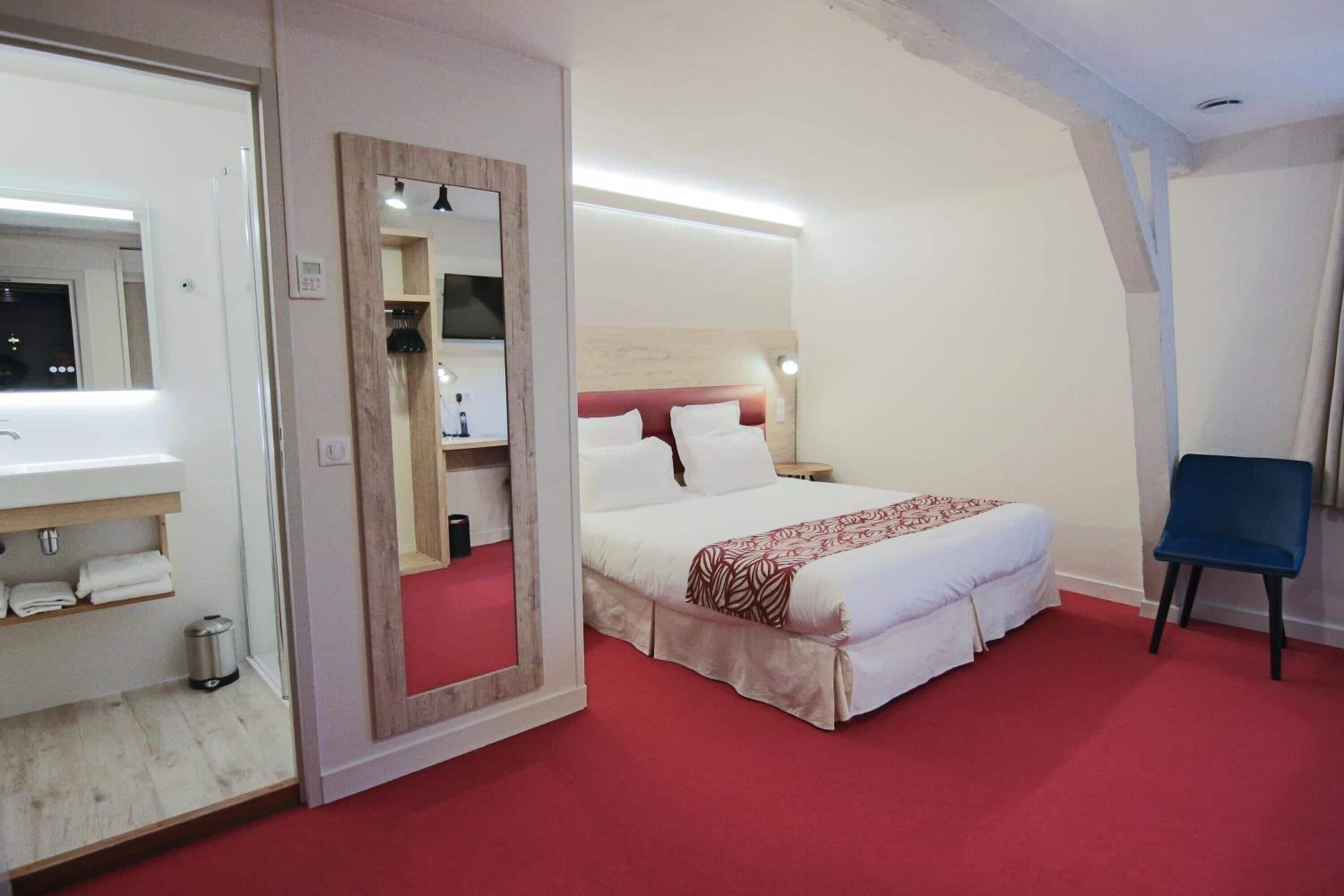 A hotel room with a bed, a chair, a mirror, and a bathroom with a sink and towel rack.