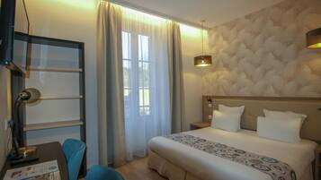 Ein Hotelzimmer mit einem Bett, einem Schreibtisch mit Lampe, einem Sessel und einem Fenster mit Vorhängen.