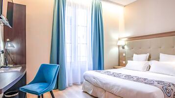 A hotel room with a bed, a desk with a chair, a lamp, and a window with curtains.