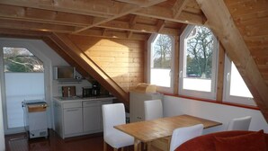 Une chambre confortable située sous les combles, avec un plafond en bois, une table à manger en bois, des chaises blanches, un canapé rouge et un coin cuisine équipé d’une cuisinière et de placards.