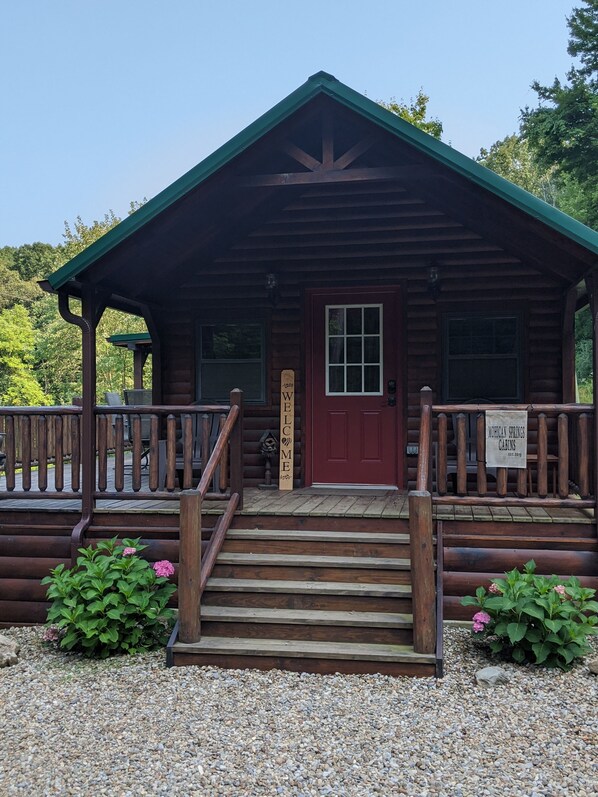A wooden cabin with a red door, a porch with a railing, and a sign that reads "Welcome".