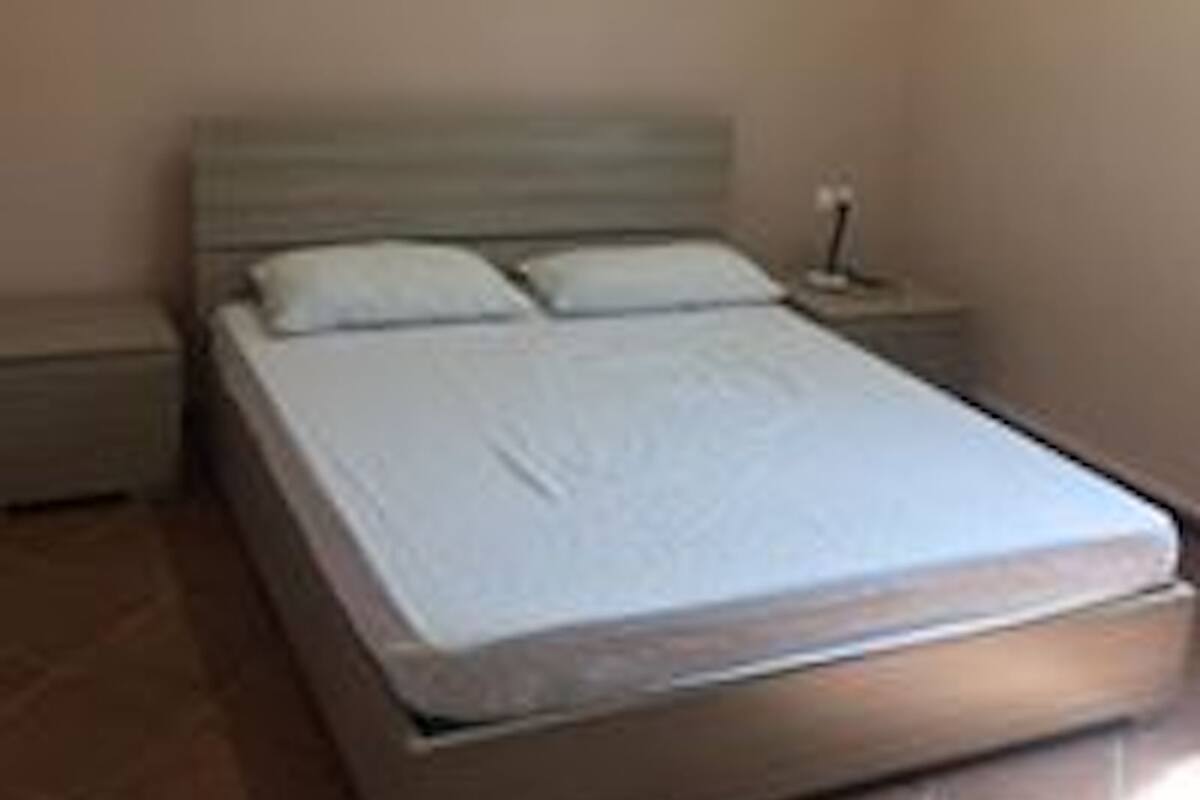 A single bed with a wooden frame and a white mattress in a room with plain walls and a tiled floor.