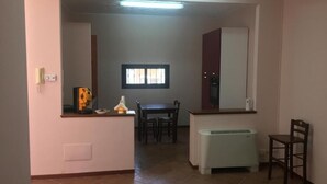 A room with a dining table, chairs, a small kitchen area with a refrigerator, and a window.