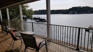 A covered deck with a table and chairs, overlooking a calm lake with boats and a forested shoreline. - LAKEFRONT PADDLEBOARD KAYAK WATERMAT SWIMMING FIREPIT GRILLING SUNDECK FISHING  (Roach)