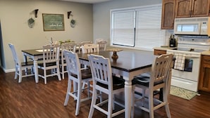 A dining area with a wooden table and white chairs, a kitchen with wooden cabinets and appliances, and a window with blinds. - LAKEFRONT PADDLEBOARD KAYAK WATERMAT SWIMMING FIREPIT GRILLING SUNDECK FISHING  (Roach)
