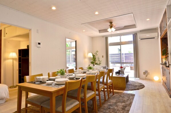 A dining area with a wooden table set for six, surrounded by chairs, with a view of the outdoors through large windows. - Purple House (Chatan)