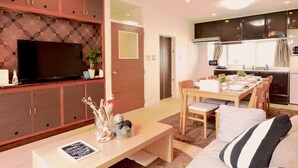 Ein modernes Wohnzimmer mit Flachbildfernseher, Holzkörperschränken, einer Essecke mit Tisch und Stühlen sowie einer Küche im Hintergrund.