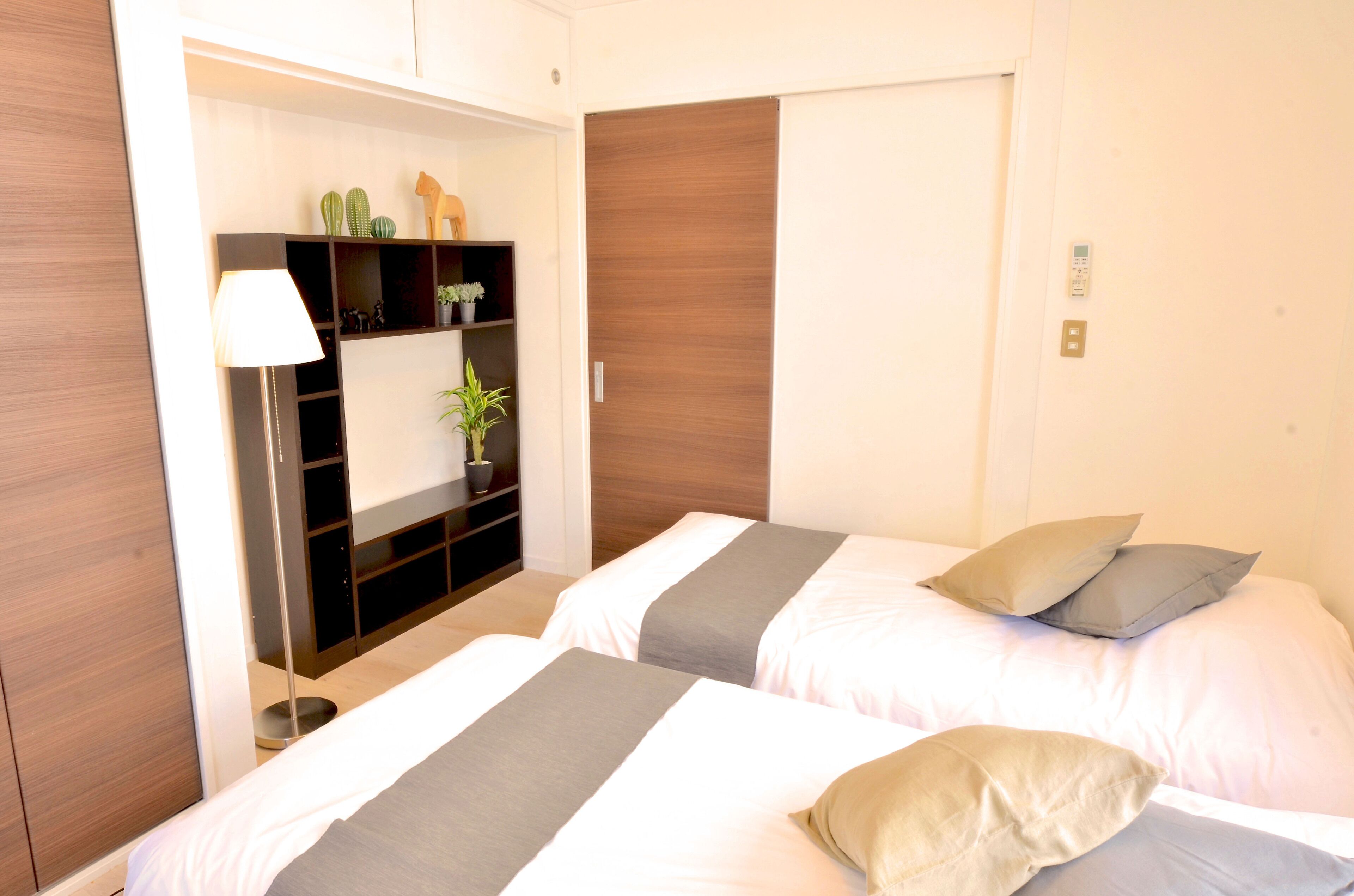 A hotel room with two beds, a bedside table, a lamp, a shelf with plants, and a wardrobe.