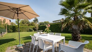 Un patio exterior con una mesa y sillas blancas, un paraguas beige y una planta en maceta.