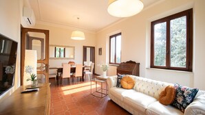 Un salón luminoso con un sofá, una mesa de comedor y sillas. Hay un televisor a la izquierda y una ventana a la derecha.