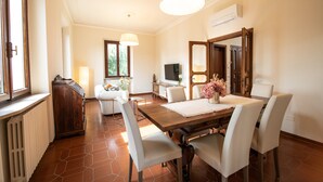 Un comedor con una mesa de madera, sillas blancas y un espejo grande. Se observa un televisor al fondo y una ventana con vistas a zonas verdes.