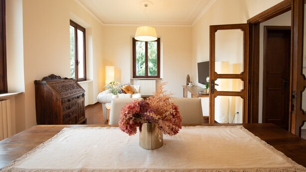 Una habitación con una mesa de madera, mantel de encaje blanco, un jarrón con flores, un sofá, una lámpara y un aparador de madera.