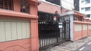 A gated entrance with a metal gate and a sign, a paved sidewalk, and a building with a pink and white facade. - Maya Service Apt (Dhaka)