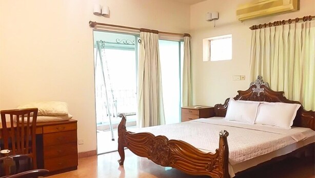 A bedroom with a wooden bed, a nightstand, a chair, and a desk. There is a window with curtains, an air conditioning unit, and a door leading outside. - Maya Service Apt (Dhaka)