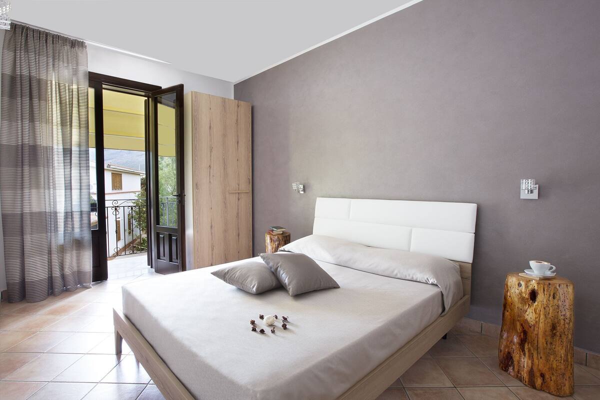 Une chambre à coucher moderne avec un lit, des rideaux, une armoire en bois, une table de chevet et un petit tabouret.